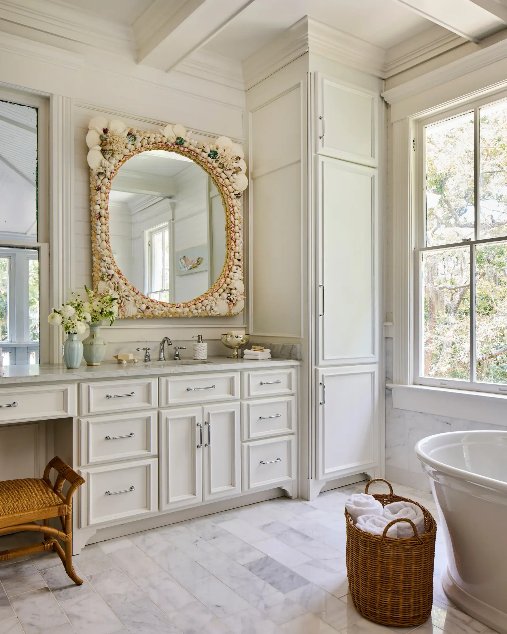 A bathroom vanity in marble beneath a large mirror framed entirely in shells, with a rattan stool at the make-up counter.