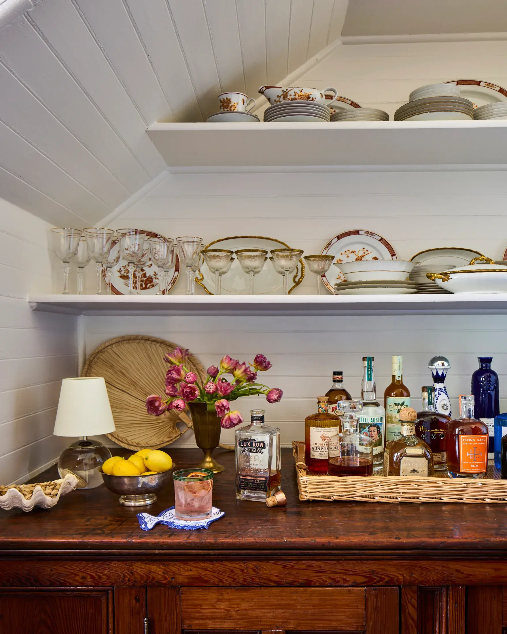 A butler's pantry under the eaves: two open shelves of gilt-edged china and coupe glasses above an antique wooden counter set out as a bar.
