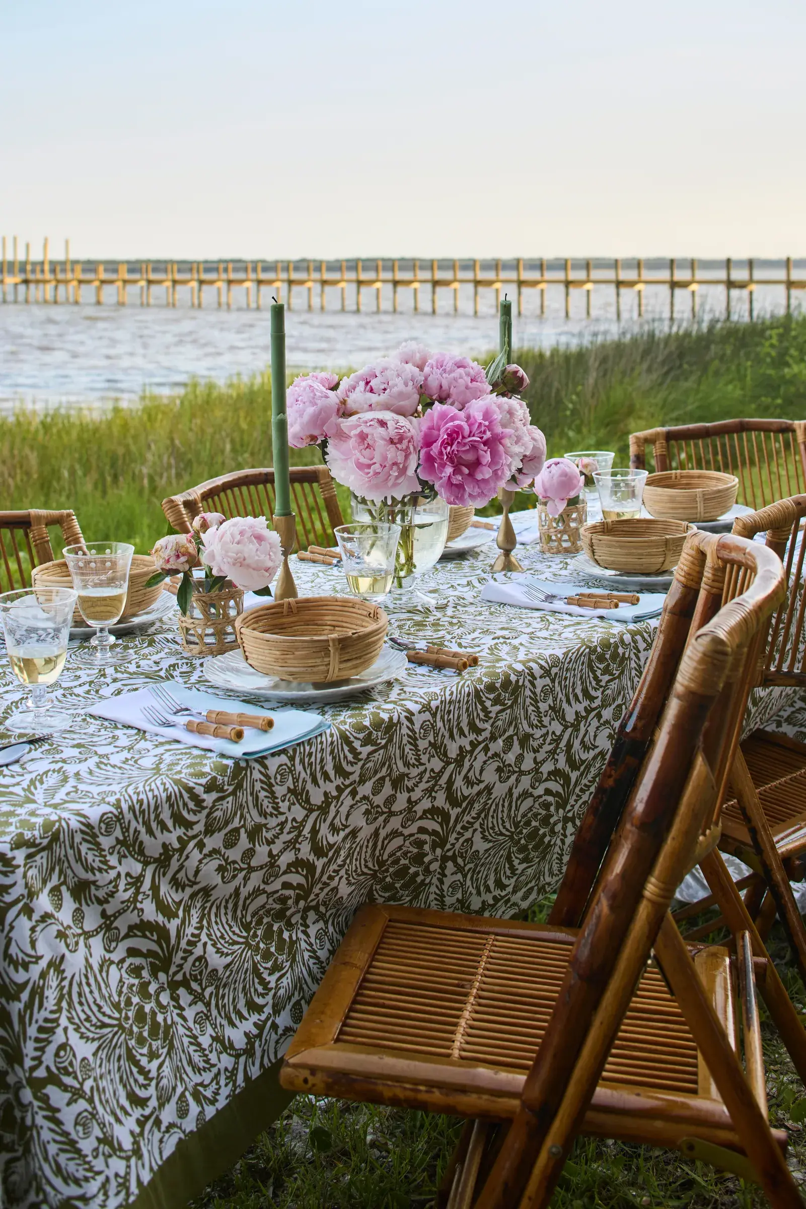 A long table laid in a green-and-white cloth at the edge of the marsh, set with peonies, rattan bowls and bamboo chairs, the dock behind it.