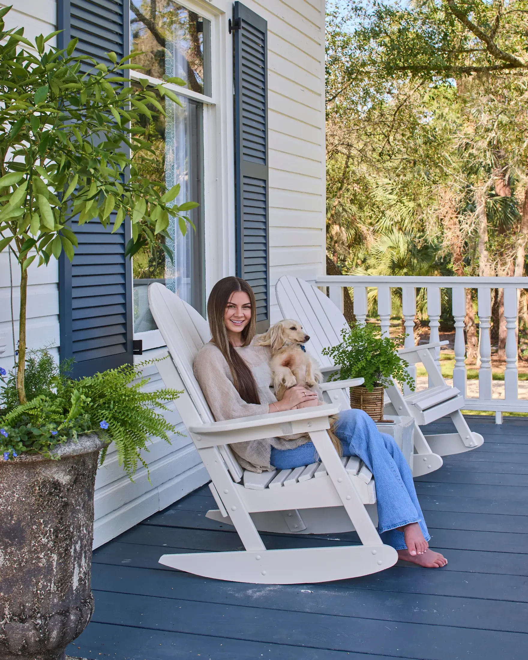 Megan McGill in a rocking chair on the front porch of the Barefield House with a dog on her lap, ferns in stone urns beside her.