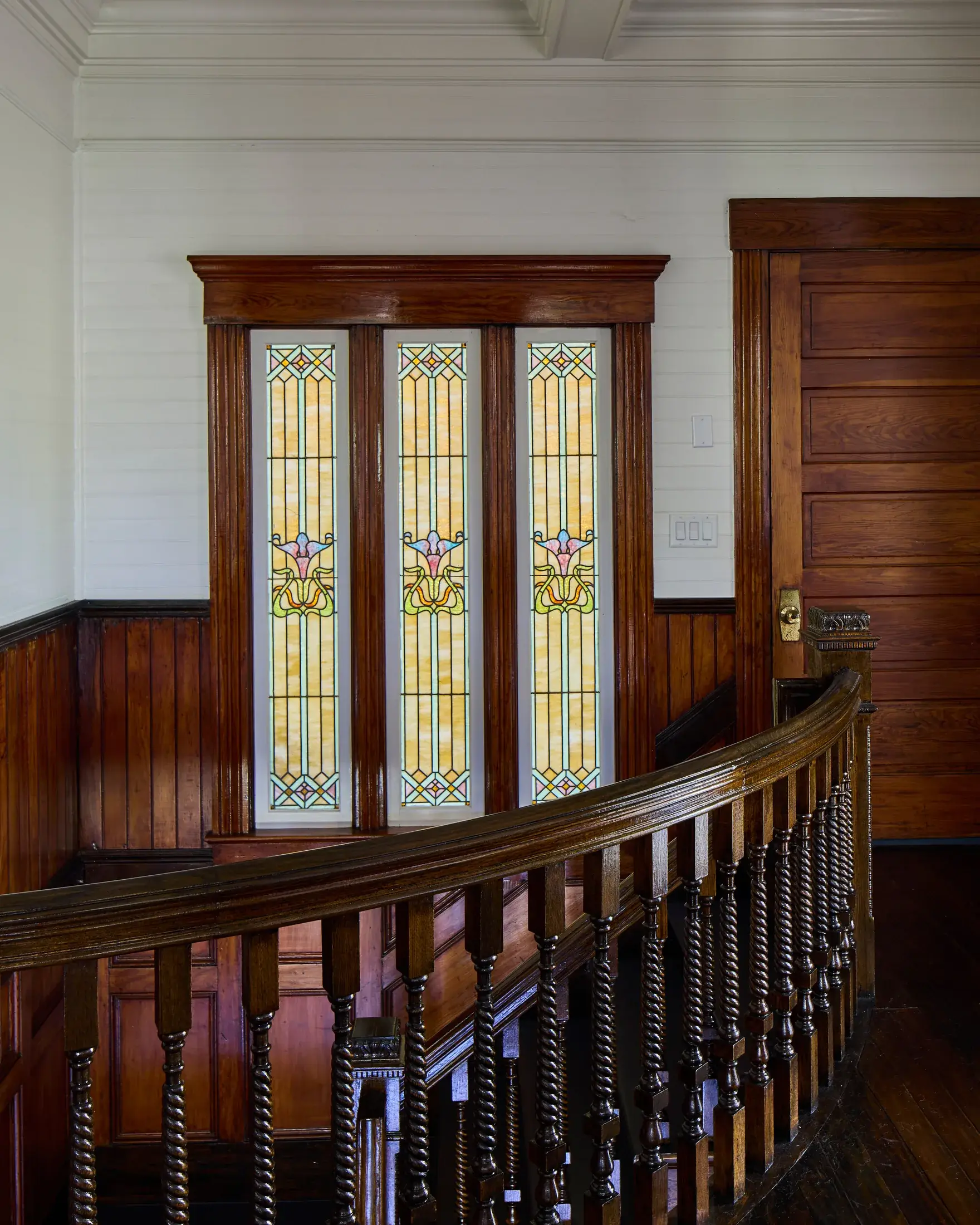 Three tall Art Nouveau stained-glass panels in amber, green and rose above the turned balusters of the stair landing.